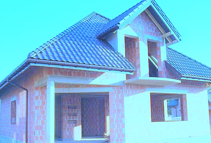 Dom w trakcie budowy z widoczną konstrukcją dachu pokrytą ciemną dachówką, ściany z czerwonej cegły i niezamontowane okna, garaż w stanie surowym otwarty.